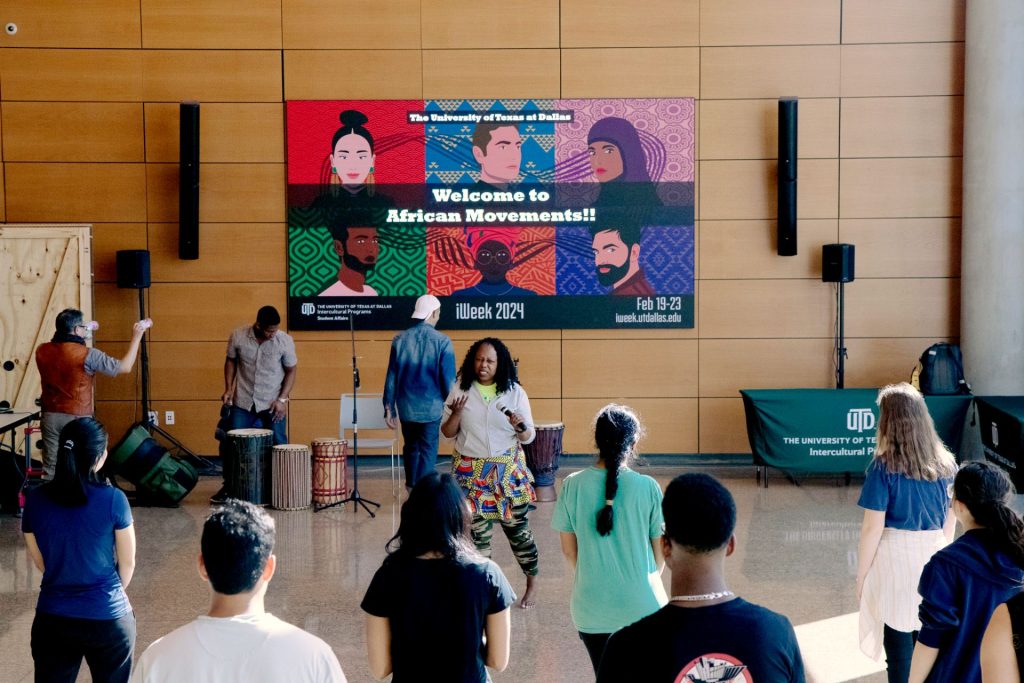 A diverse group of people gathered in front of a large screen, celebrating African dance and drumming traditions at UT Dallas.