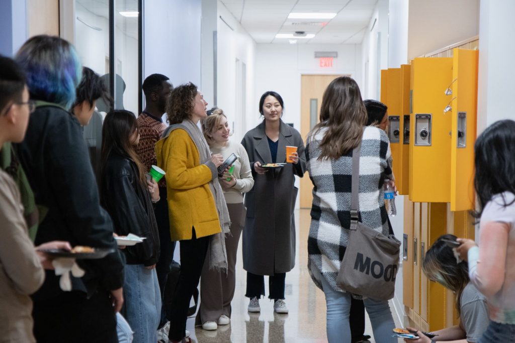 A group of students standing in a hallway with yellow lockers. This is the 2024 ATEC Pop-up Locker Exhibition at The University of Texas at Dallas, where twenty students from various disciplines showcased their artistic projects exploring themes of isolation and belonging.